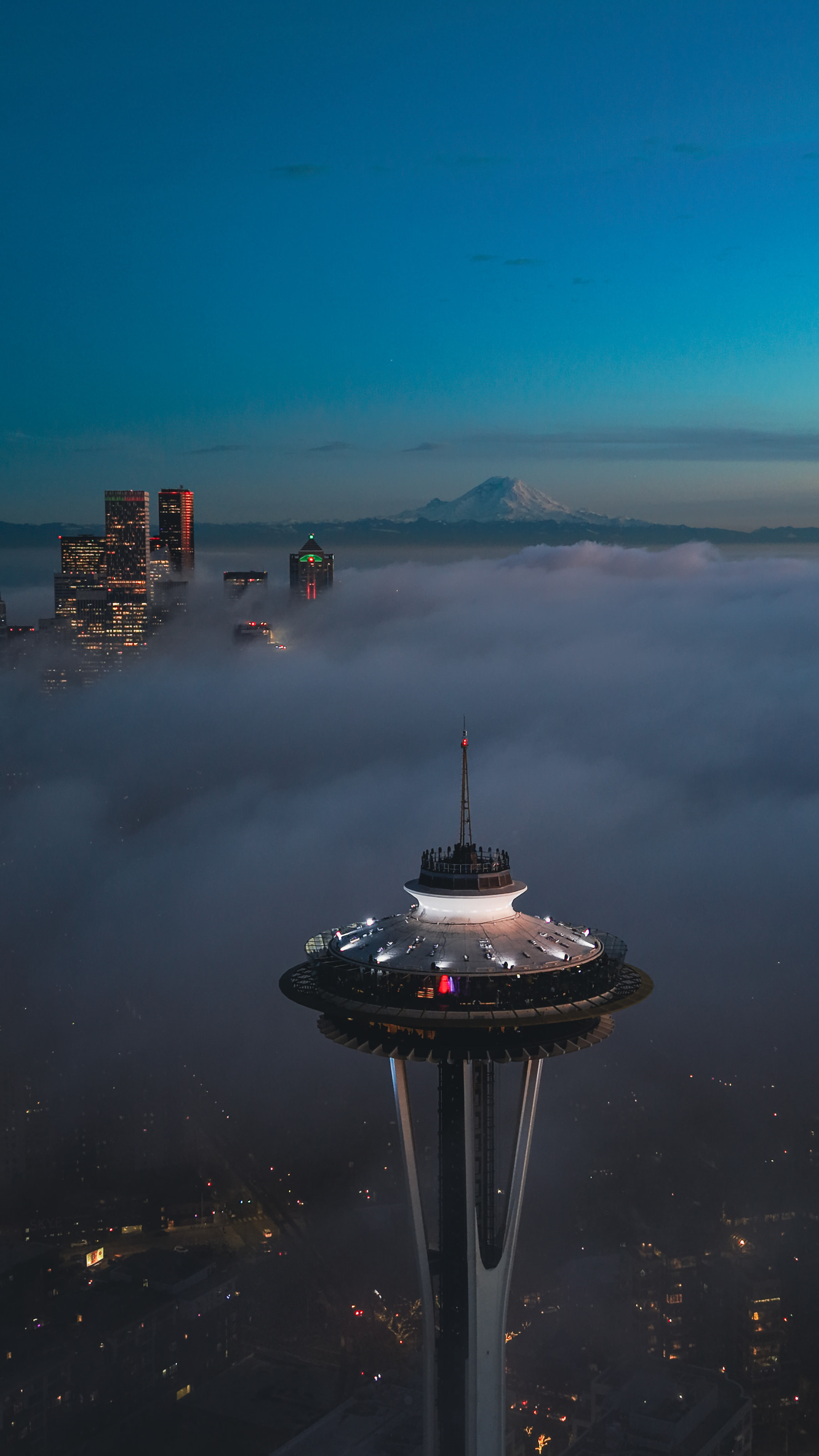 Aerial view of the top of the Space Needle rising from a sea of fog at blue hour, with downtown towers and Mount Rainier on the horizon.