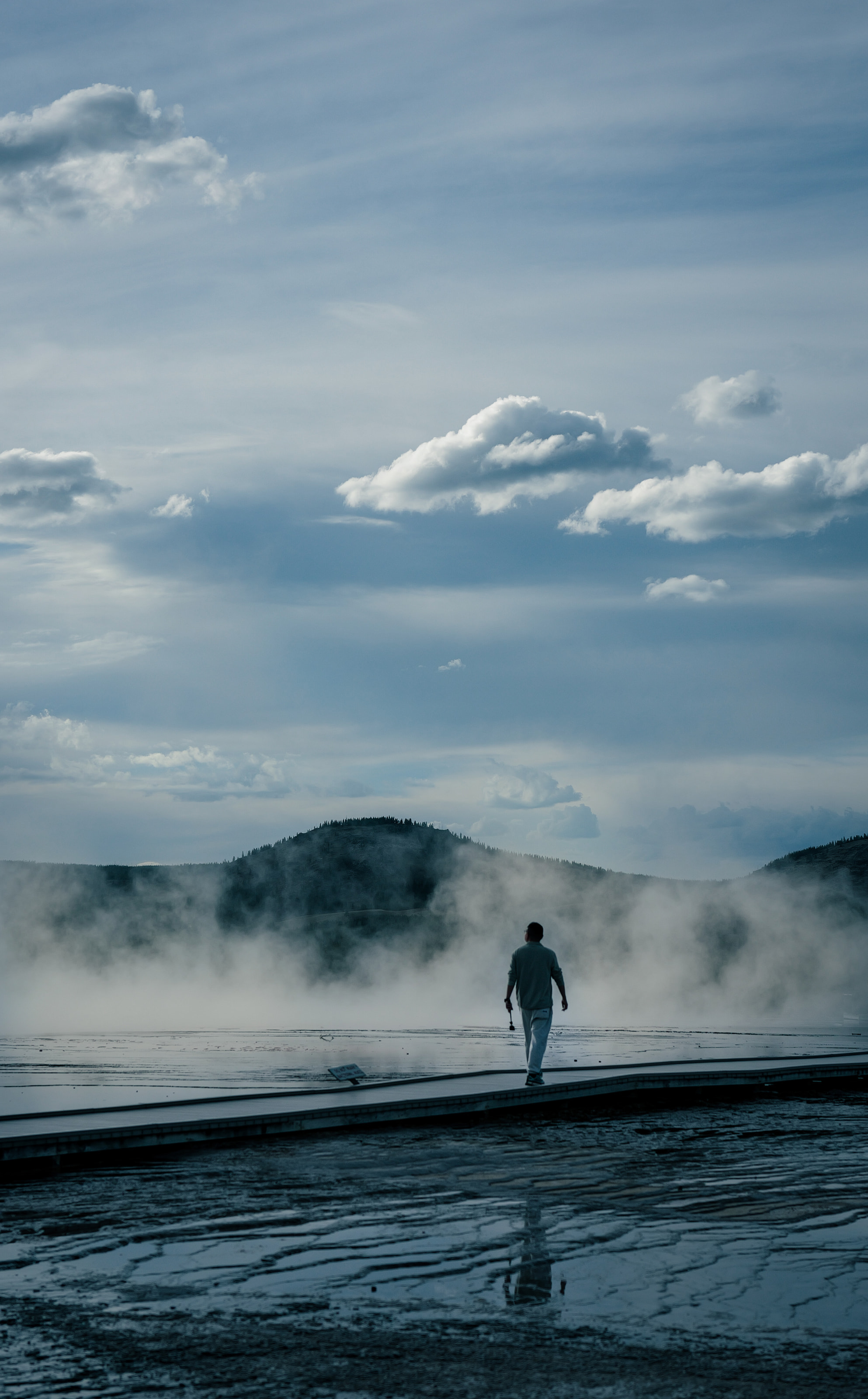 A person walking along a boardwalk toward geothermal steam under a cloud-filled sky.