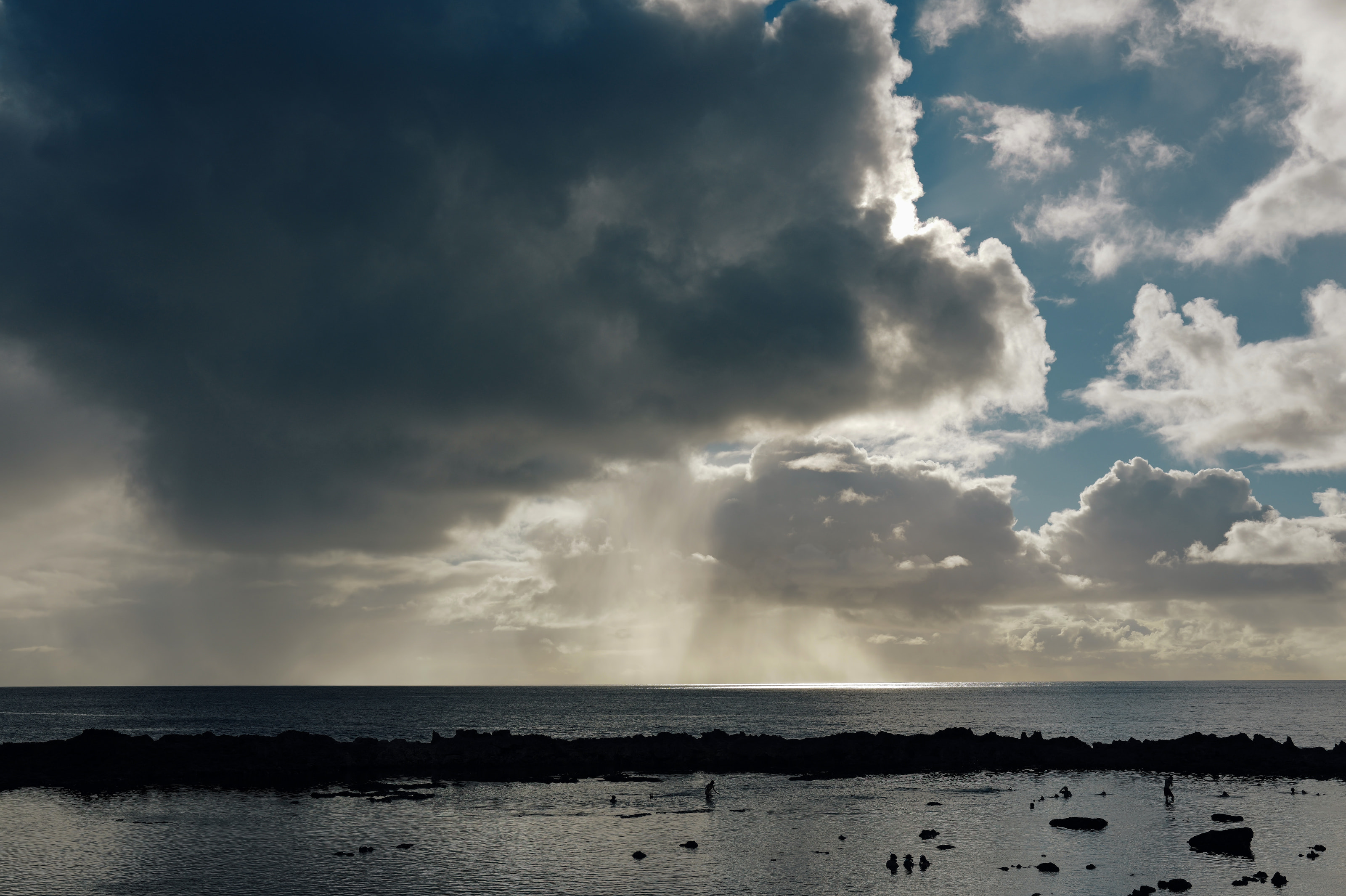 A rain squall and sun rays over the ocean, with swimmers in a tide pool below.