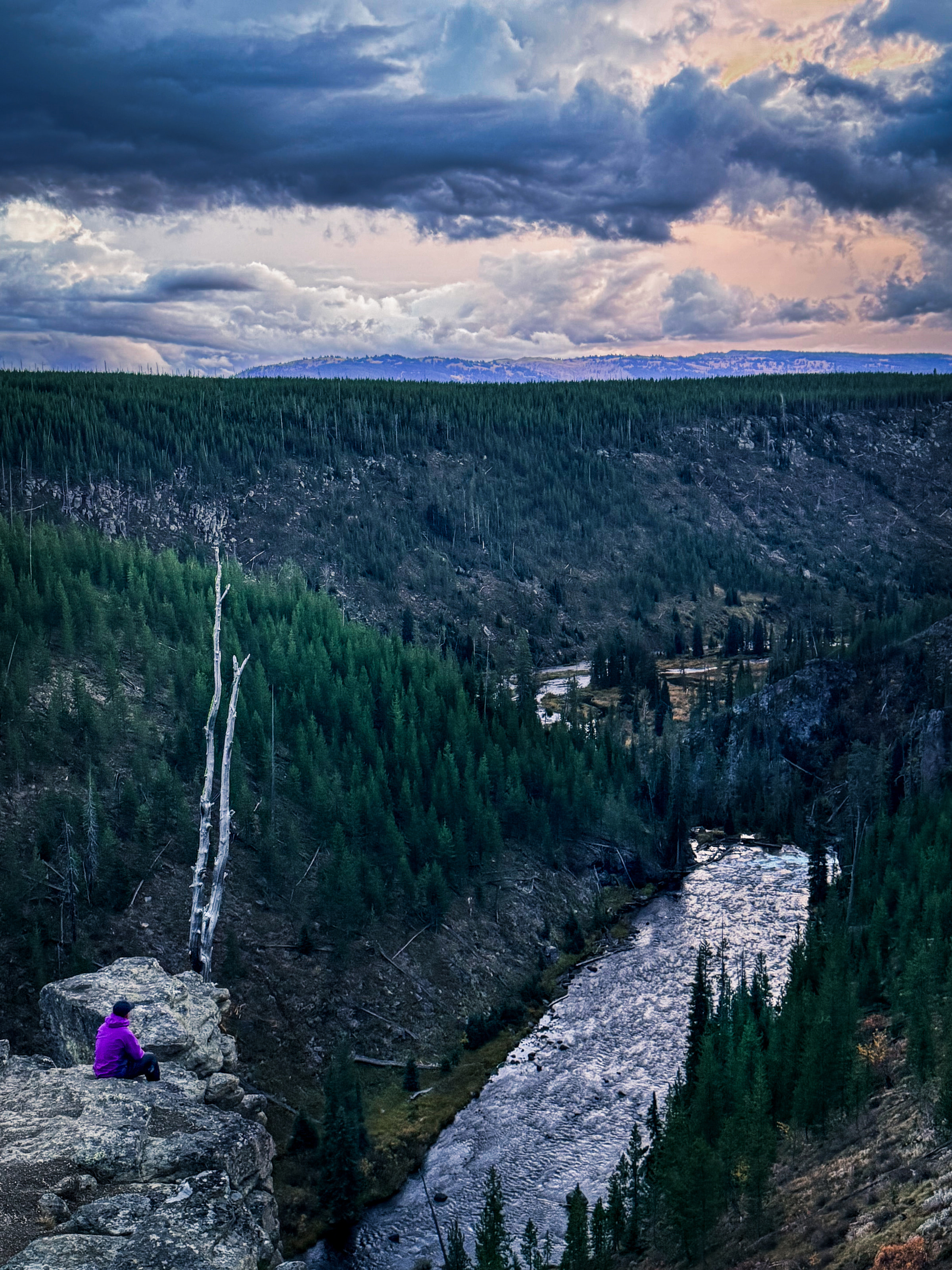 A figure in a purple jacket sitting on a cliff above a river canyon under storm light.