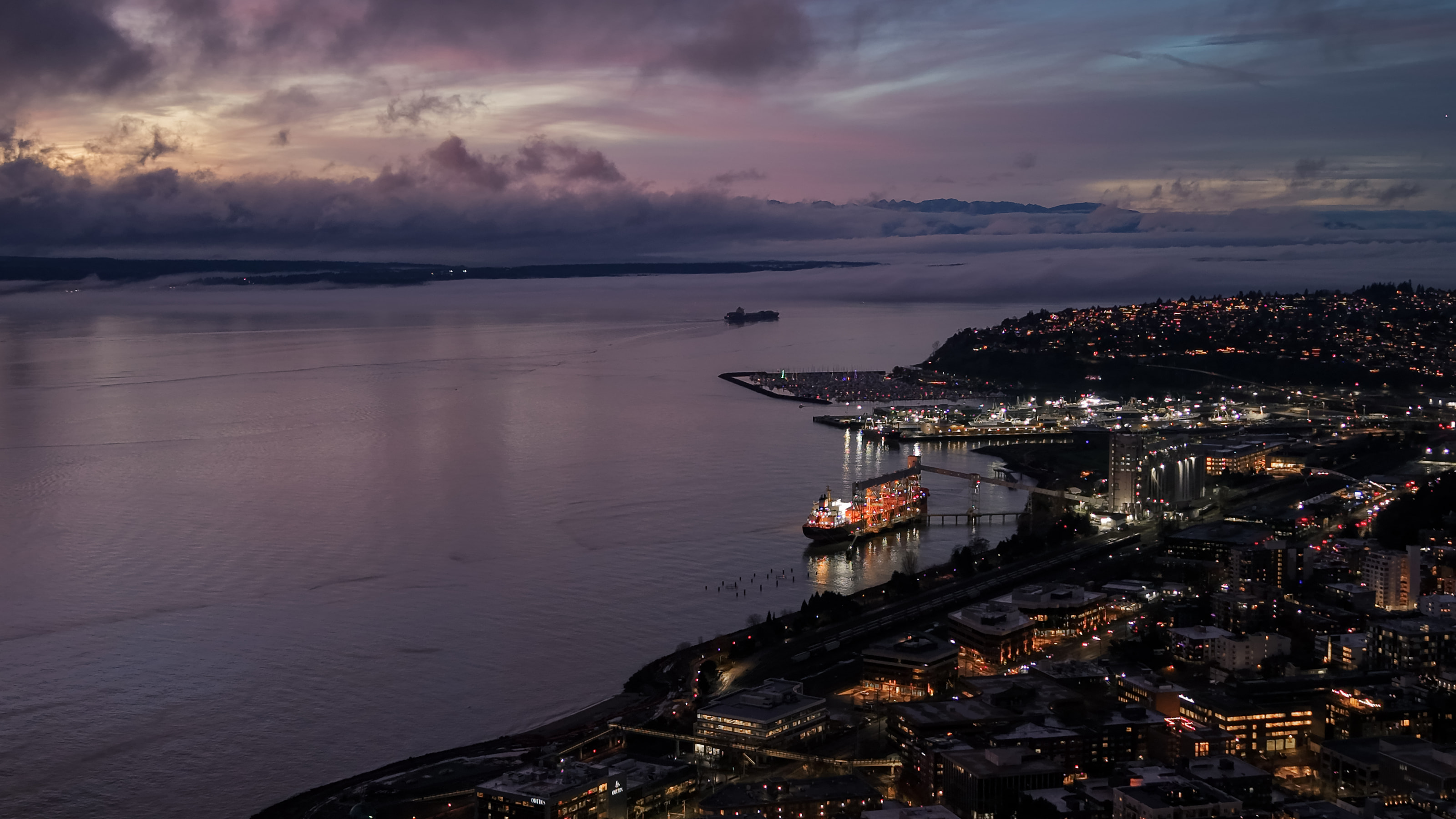 Aerial view of a waterfront at dusk, a lit freighter at the pier and a fog bank beyond.