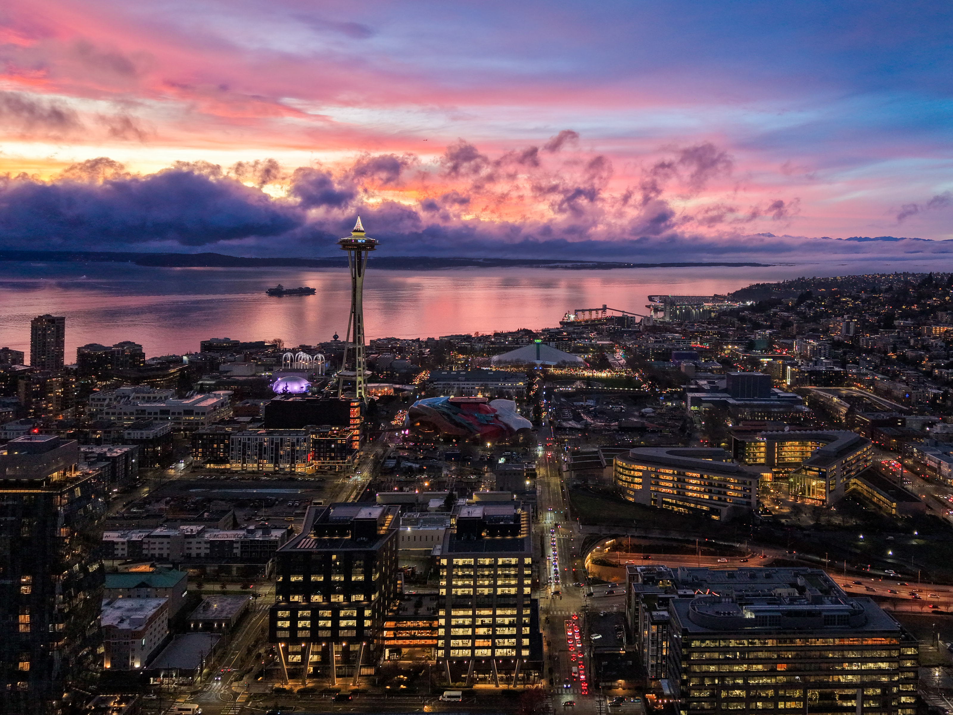Aerial view of the city at dusk under pink and violet clouds, the Space Needle and lit streets leading to the bay.