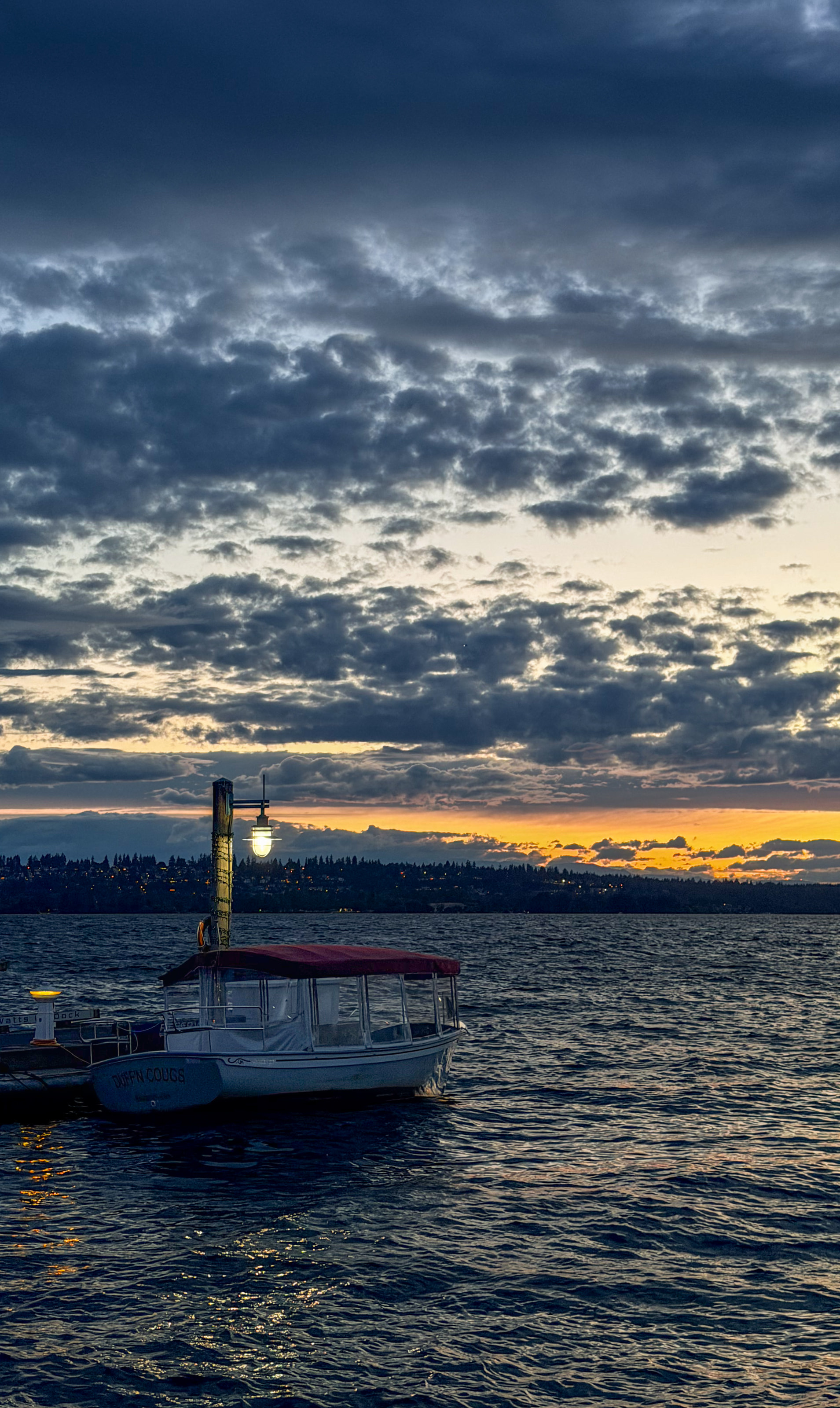 A small boat moored at a dock beside a lit lamp, under dramatic clouds at sunset.