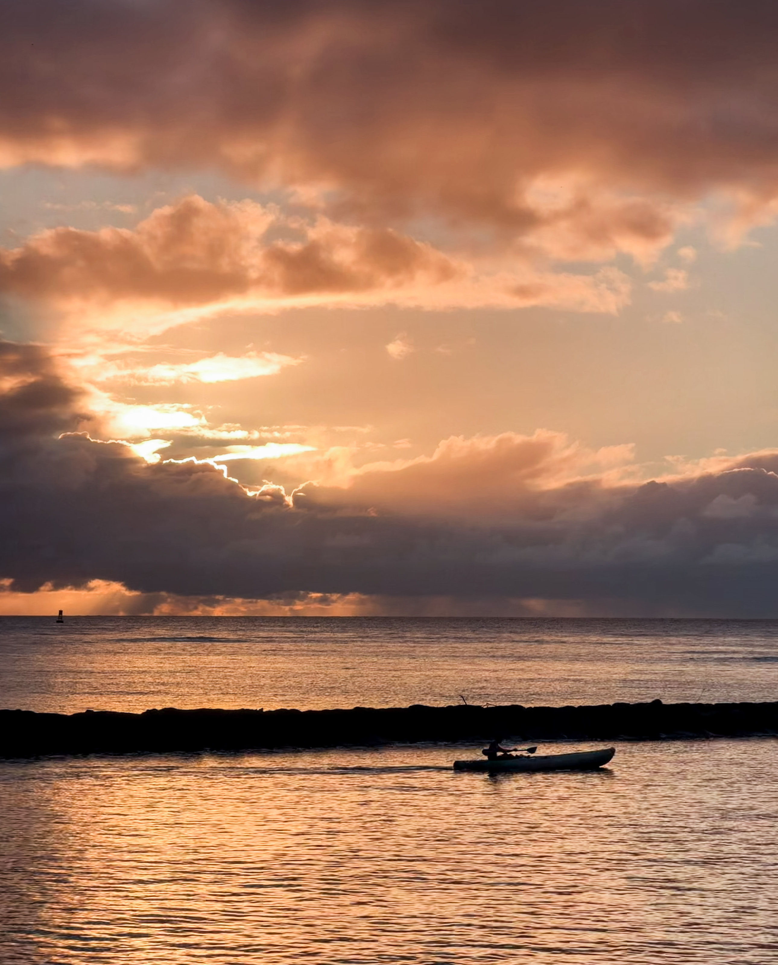 A kayaker paddling beyond a breakwater beneath peach-colored clouds.