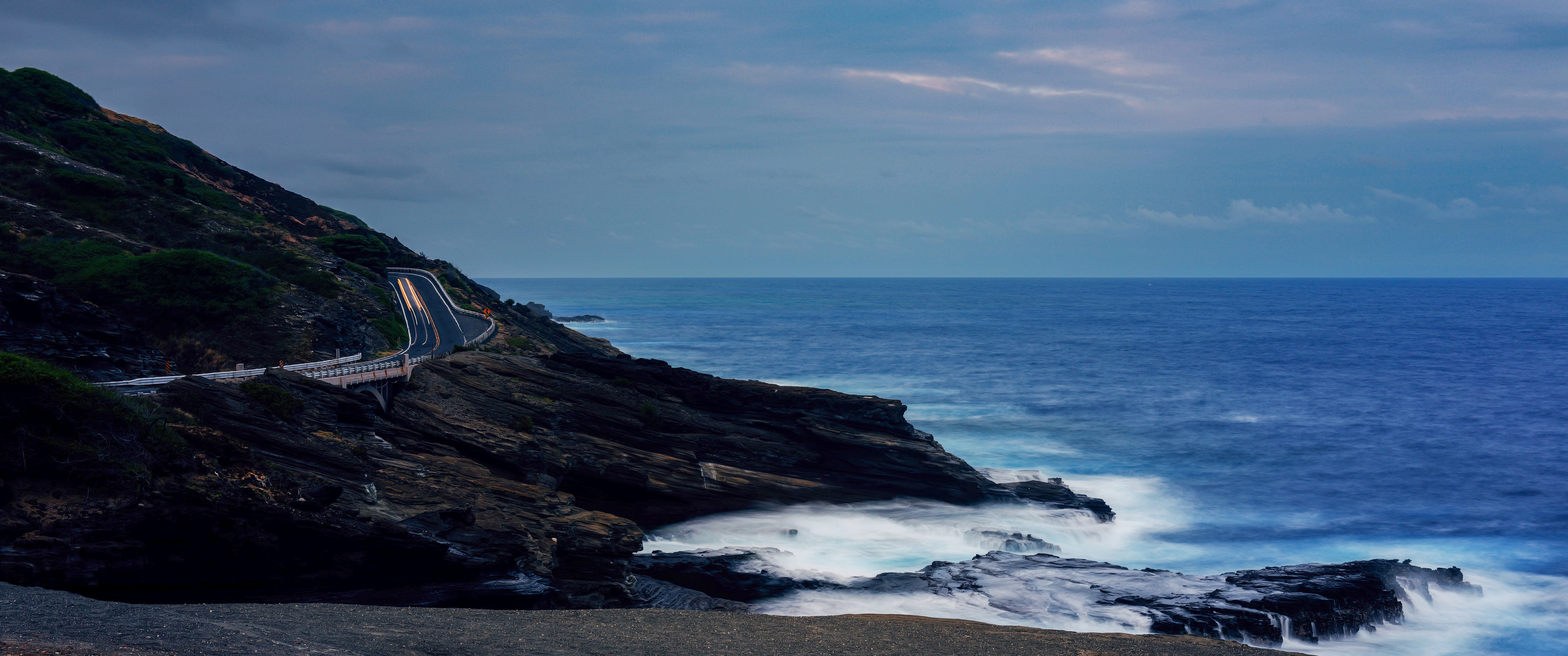 A coast road with car light trails above long-exposure surf on lava rock at dusk.