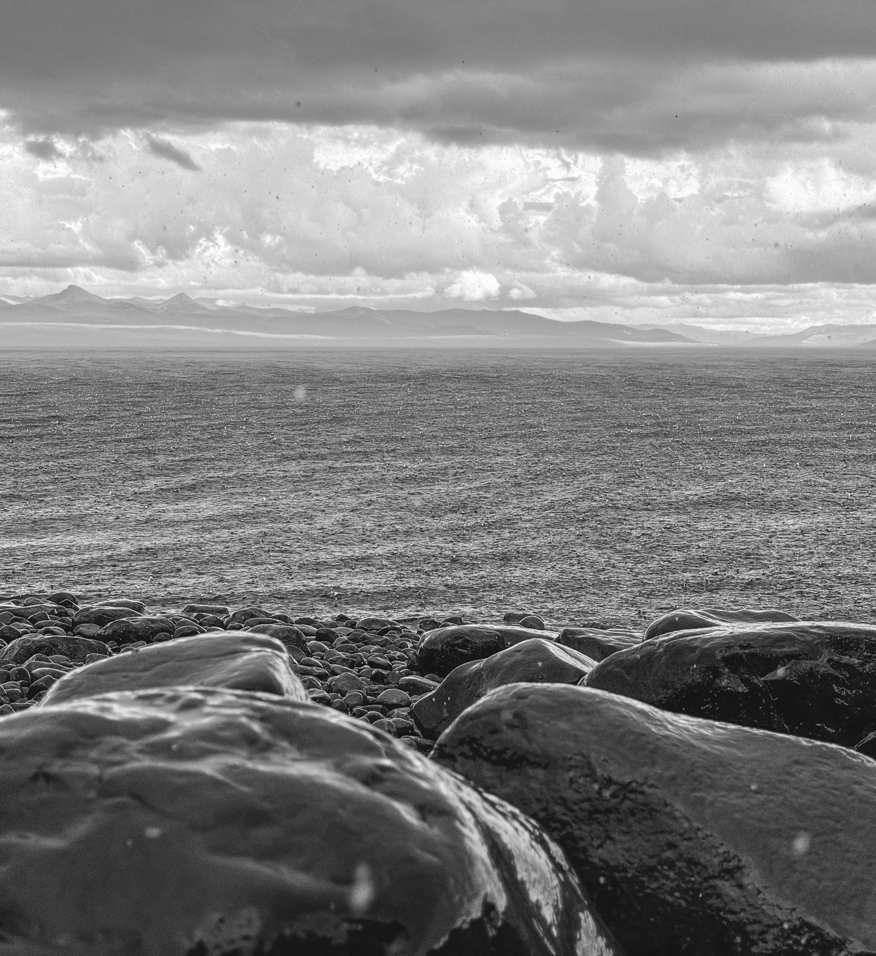 Black-and-white view of wet boulders on a shore, a lake, mountains and storm clouds.