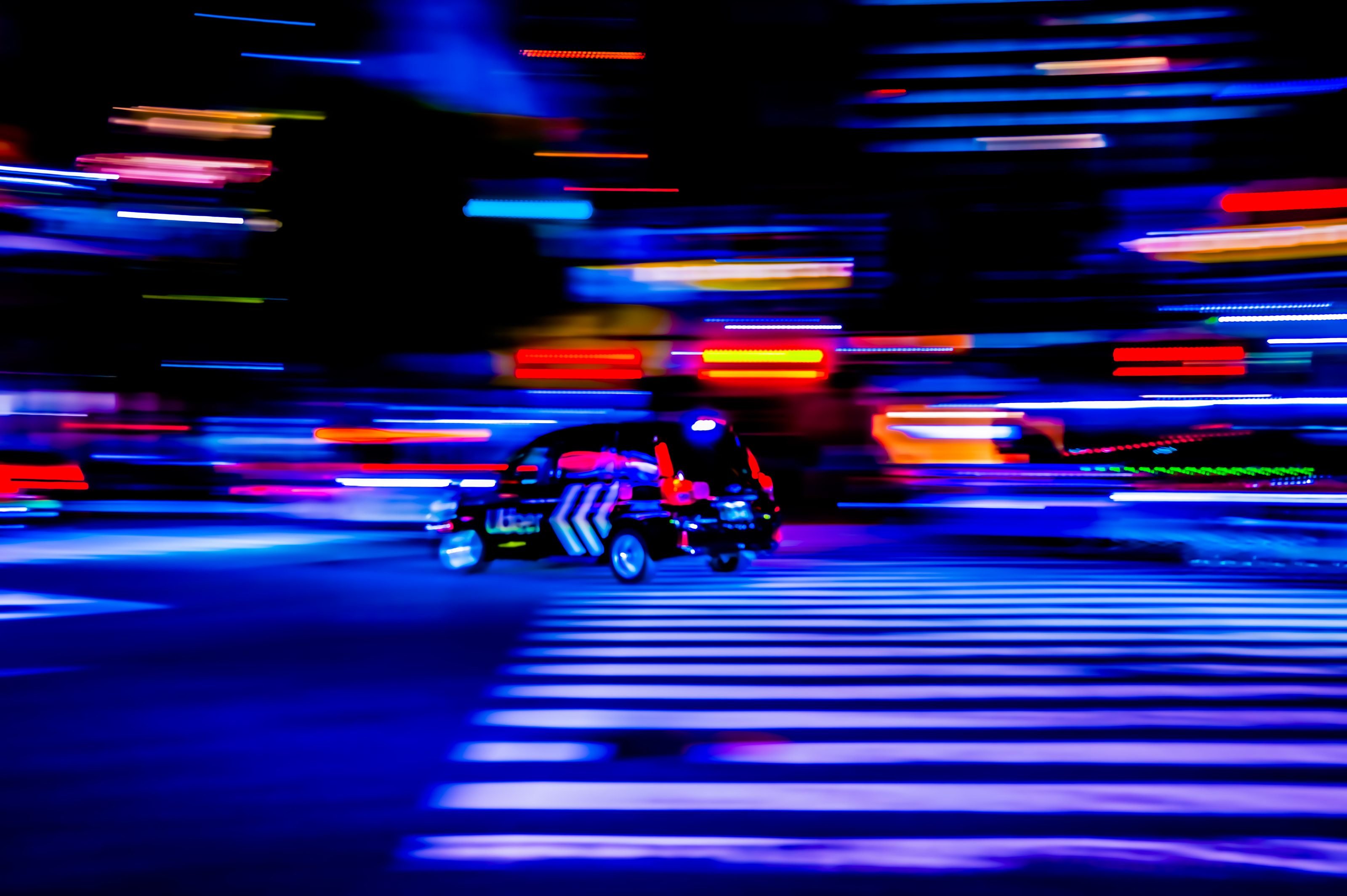 A panned photograph of a taxi crossing a crosswalk through streaks of blue and red neon.
