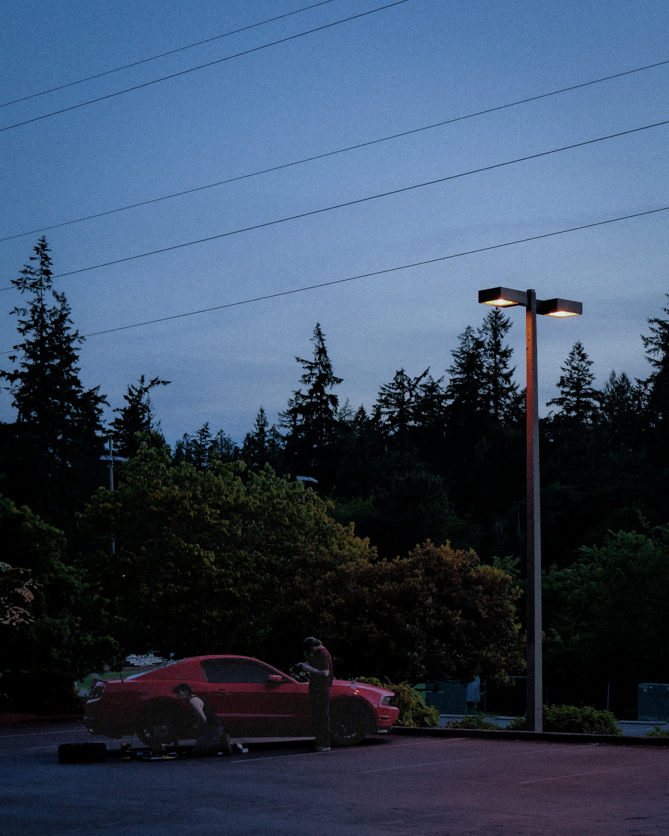 Two people working on a red car under a lone streetlight at blue hour, with fir trees and power lines above.