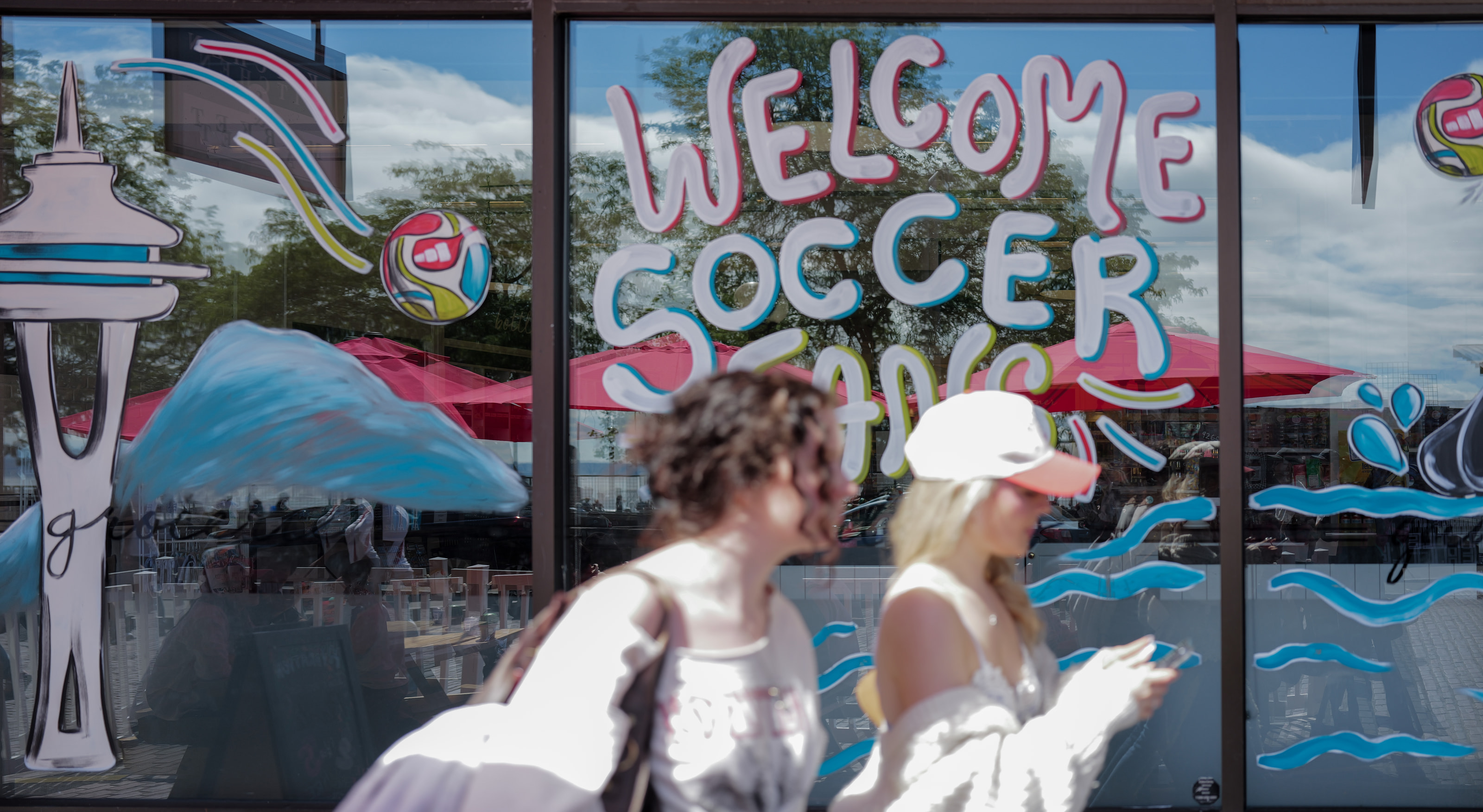 A painted shop window greeting soccer fans, with two women walking past in bright sun.