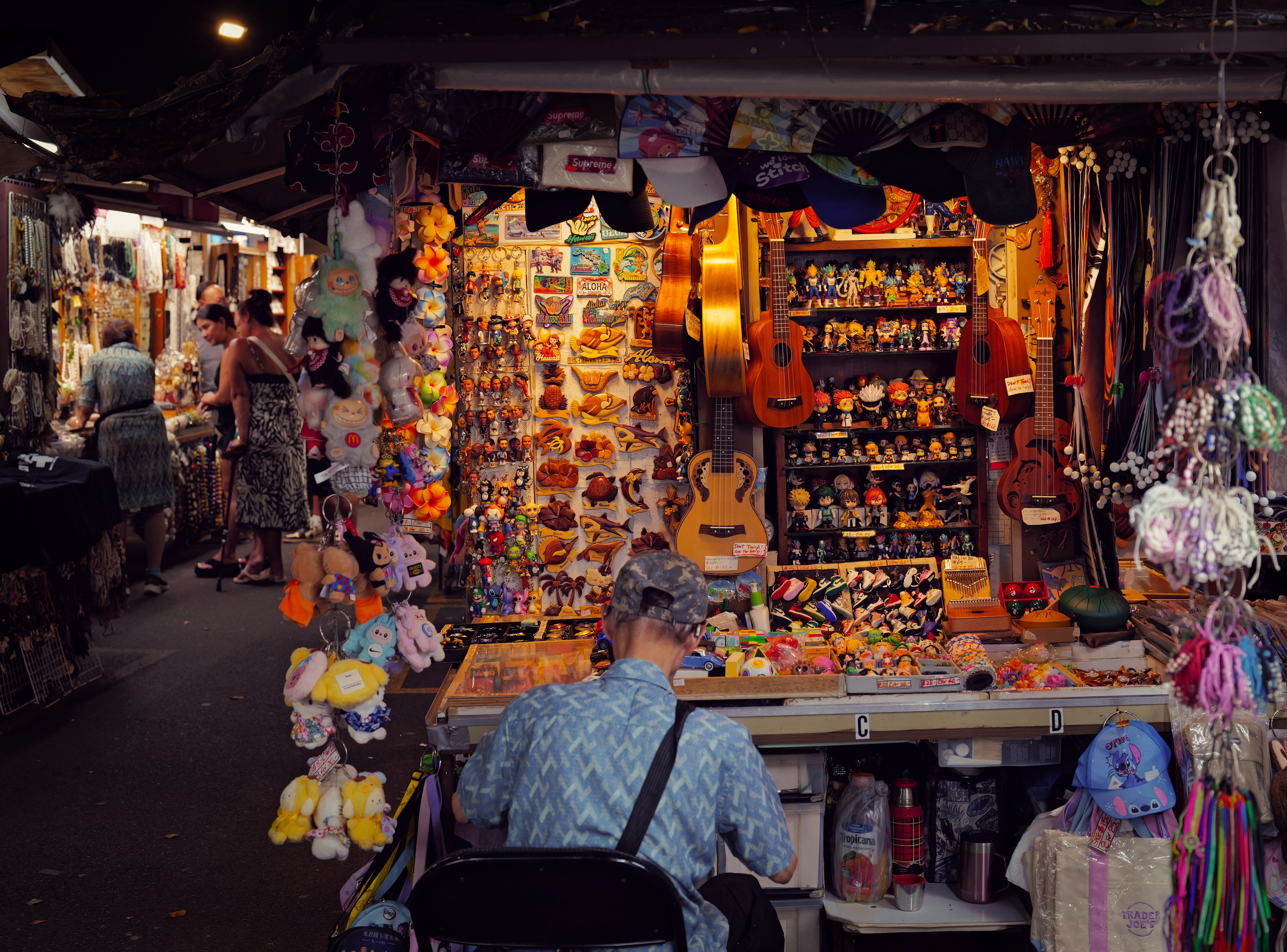 A seated vendor surrounded by ukuleles, keychains and figurines in a market stall.