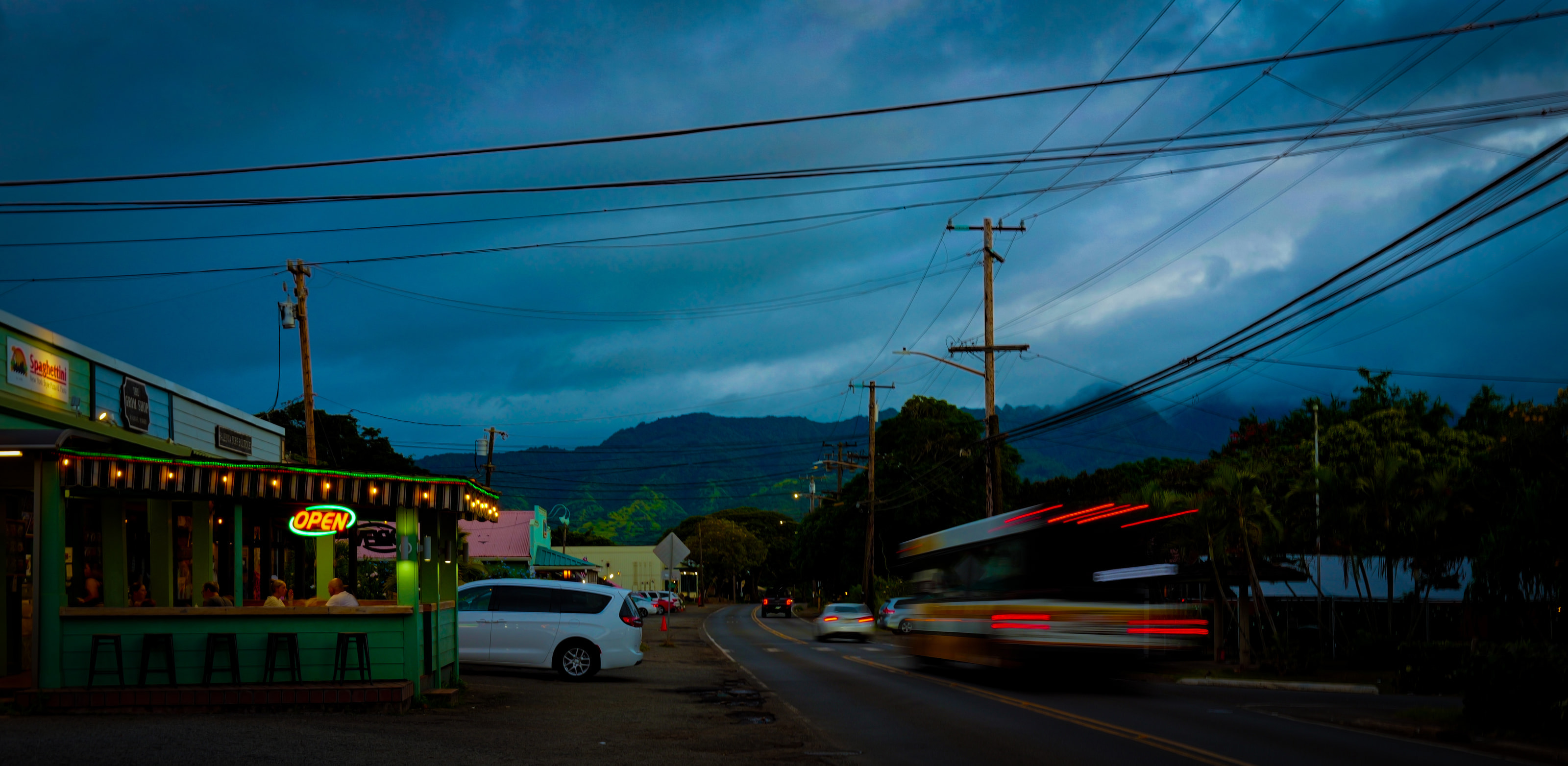 A green diner with an OPEN sign at blue hour, a bus blurring past under power lines, mountains behind.
