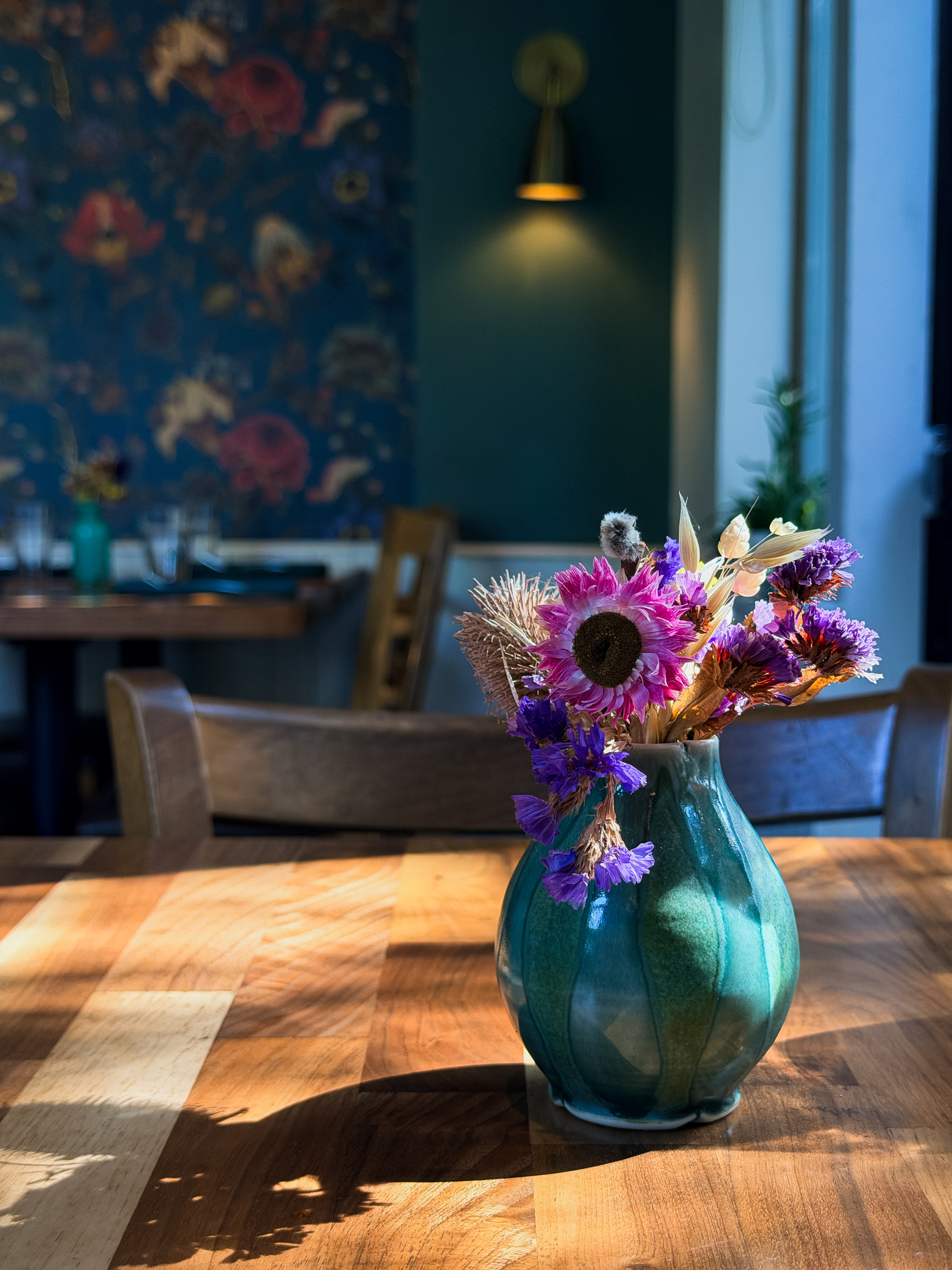 A teal vase of dried flowers on a sunlit wooden table, with floral wallpaper behind.