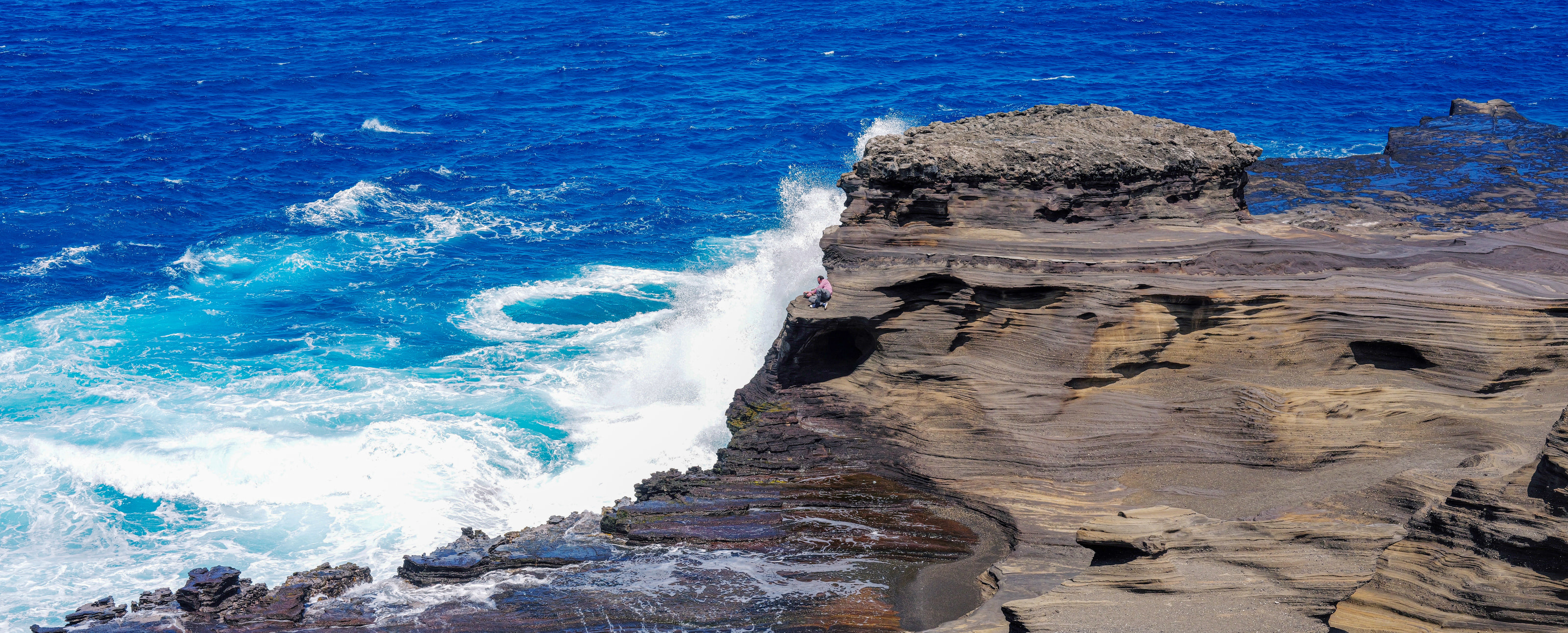 A wave breaking over a lava shelf into electric blue water, with two tiny figures on the rock.