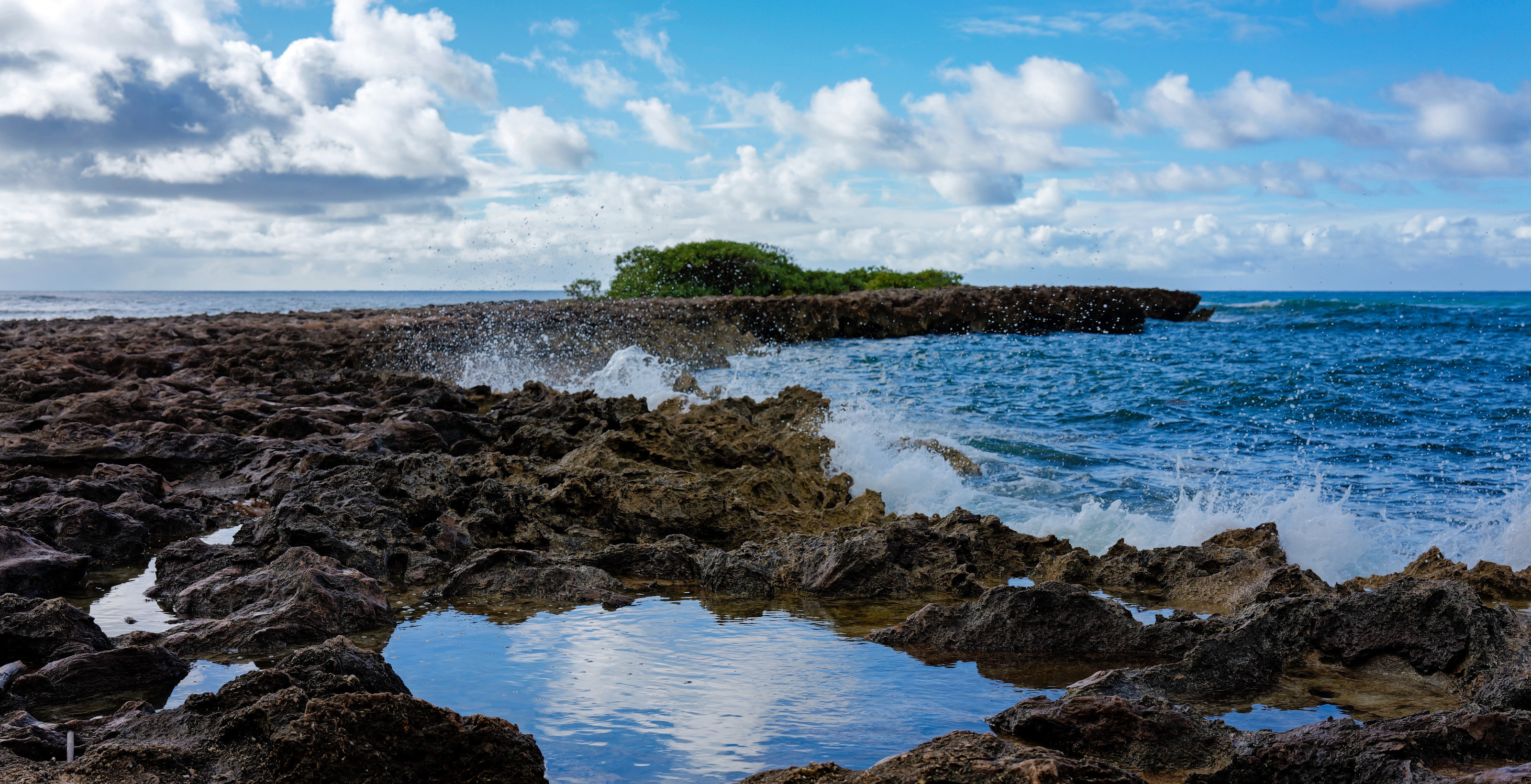 A tide pool mirroring clouds among lava rocks, with surf behind.