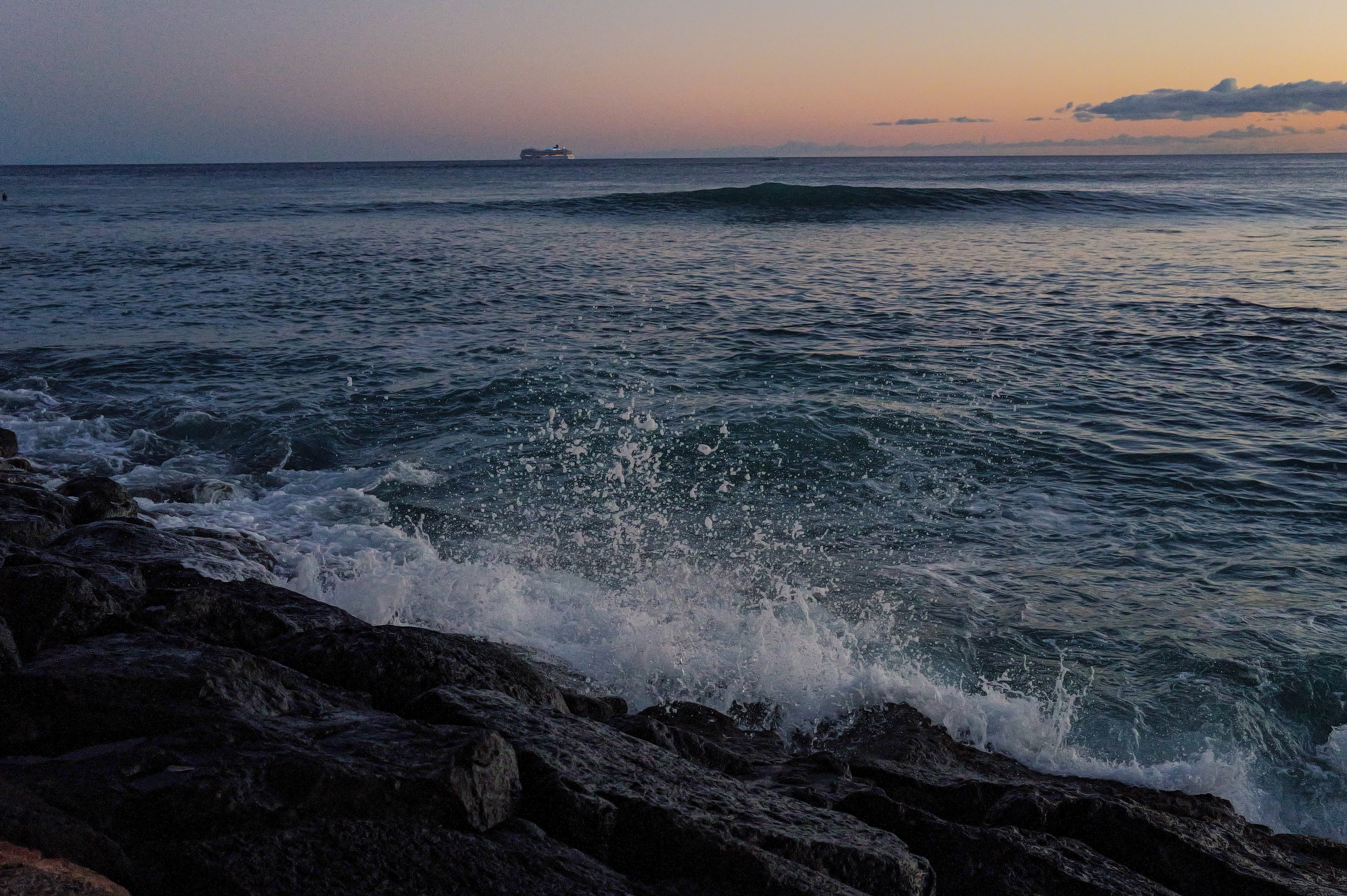 Surf bursting over dark rocks at dusk, with a cruise ship on the horizon.