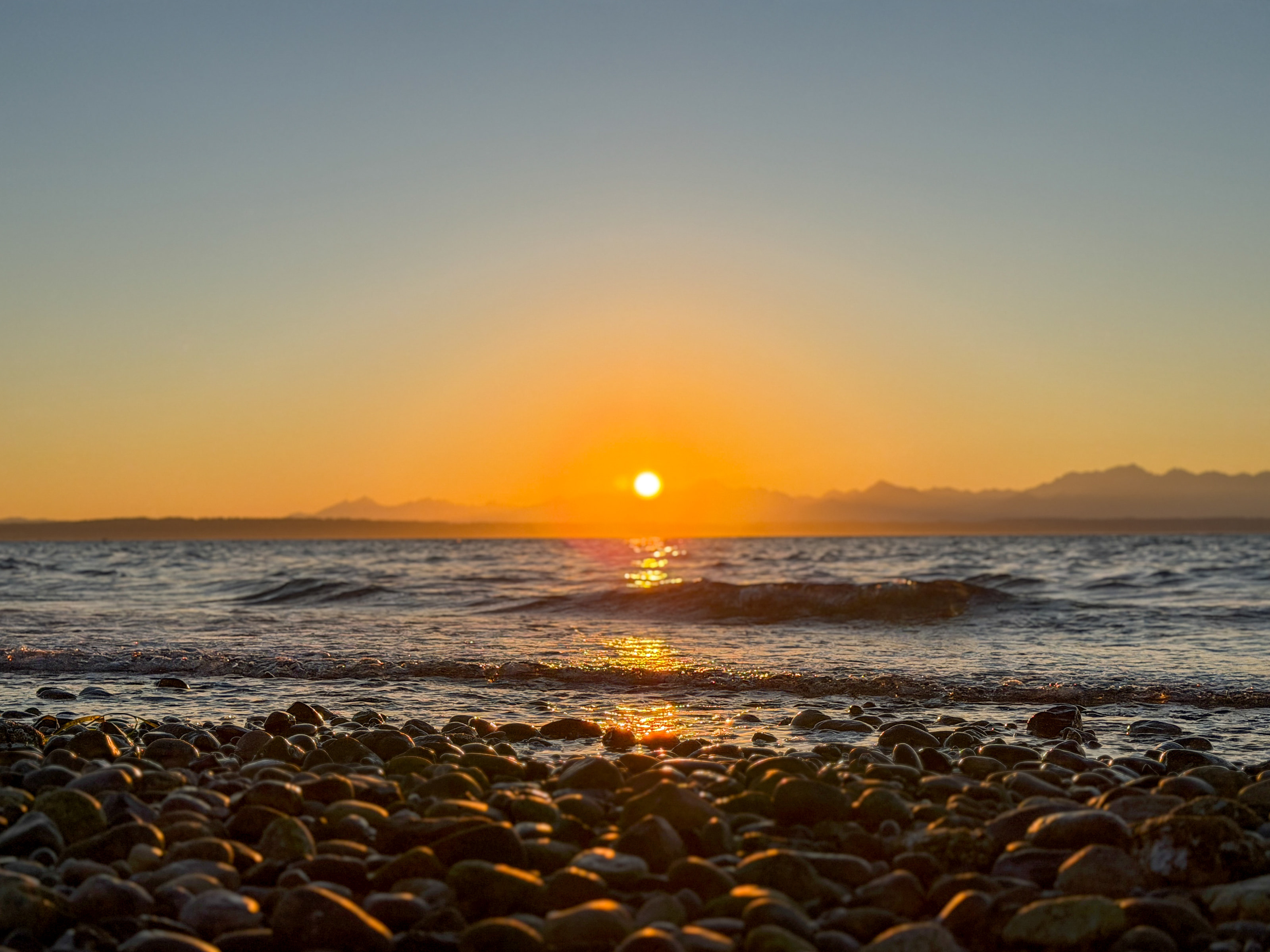 A wet pebble beach with the sun touching a mountain horizon.