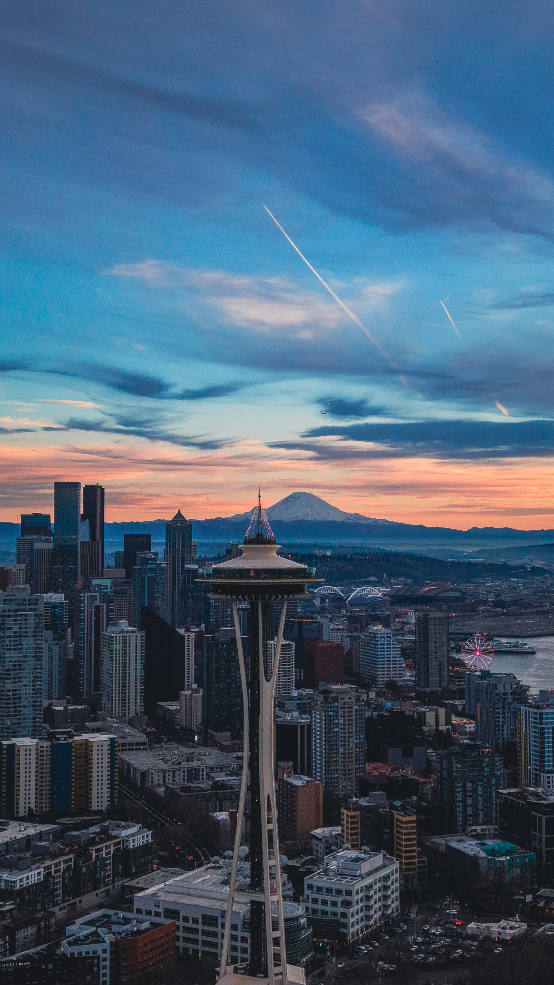 Aerial view of the Space Needle with Mount Rainier and contrails at sunset, a Ferris wheel on the waterfront.